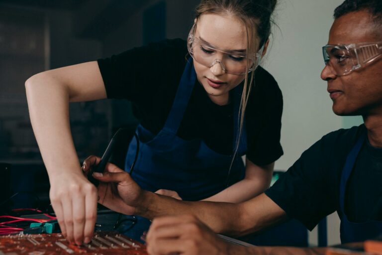 Two engineers wearing protective gear work together on a circuit board, focusing on technical precision.