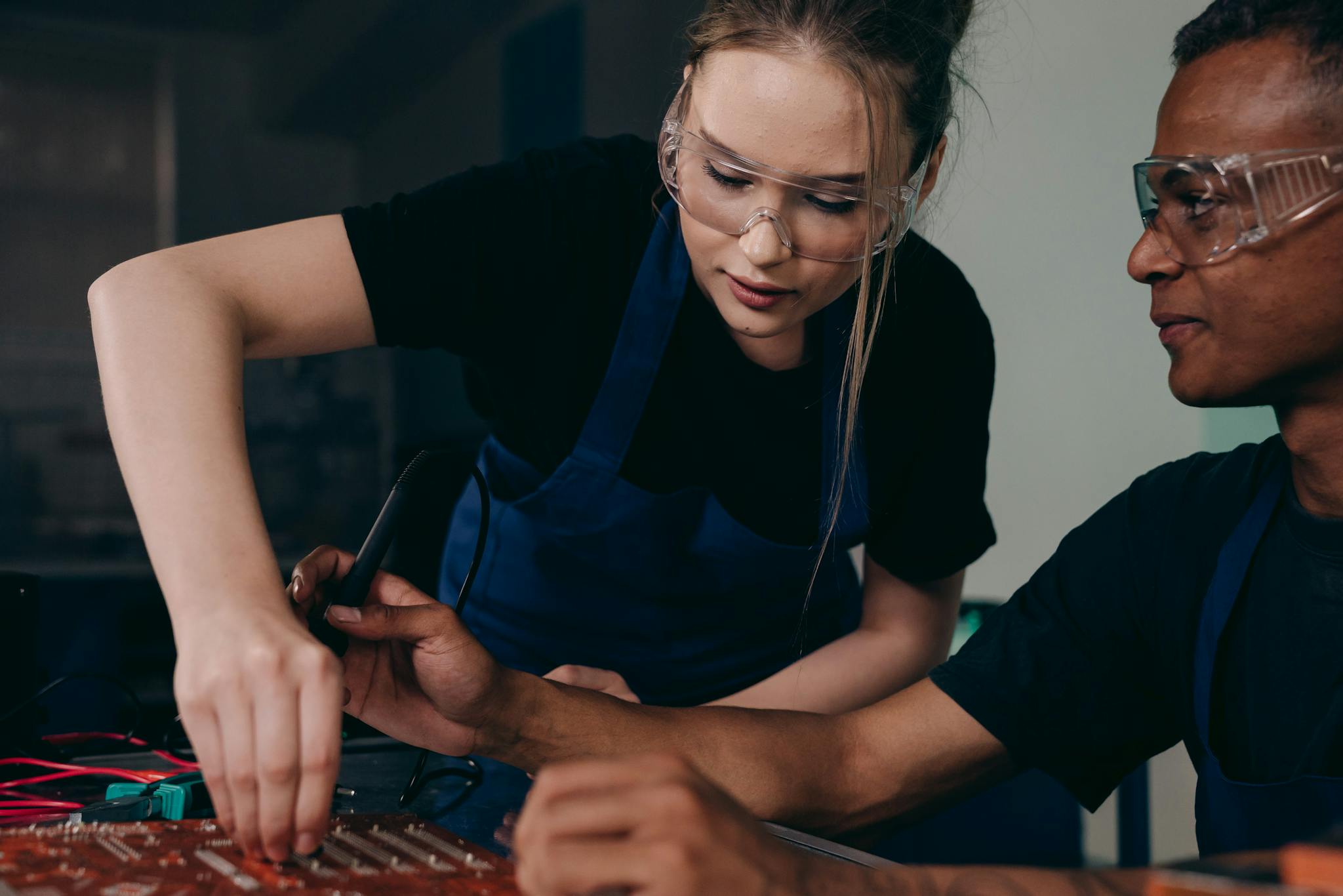 Two engineers wearing protective gear work together on a circuit board, focusing on technical precision.